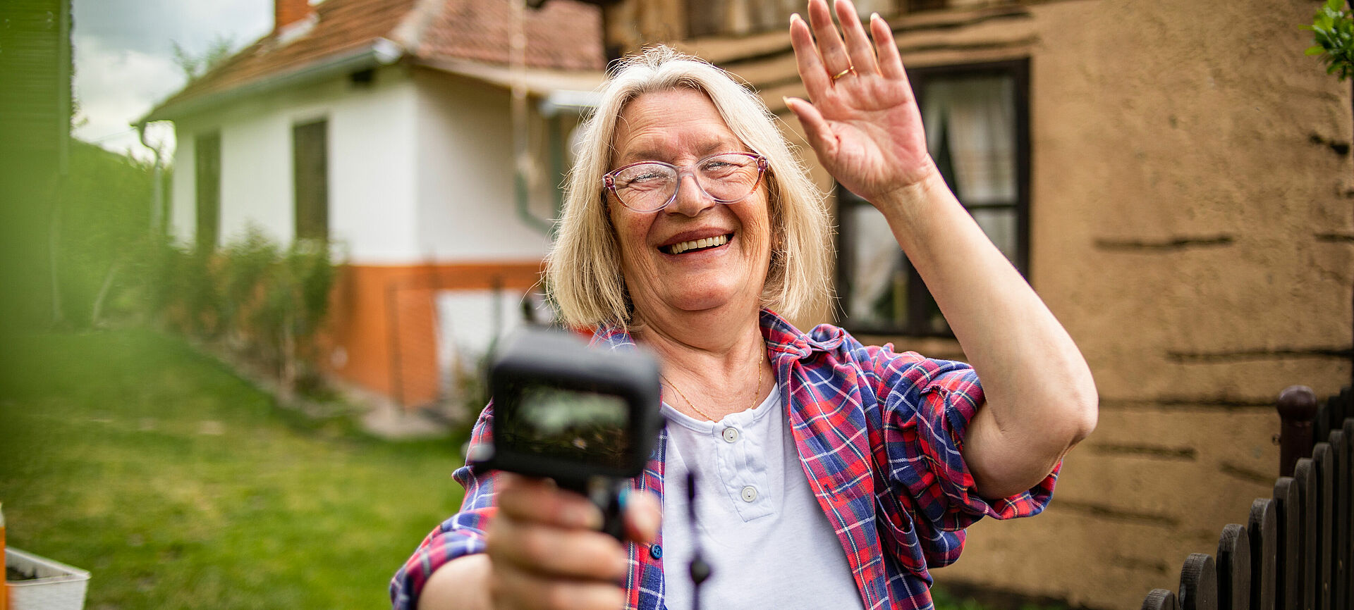 Eine Frau, die in ihrem Garten lachend in eine Kamera winkt.