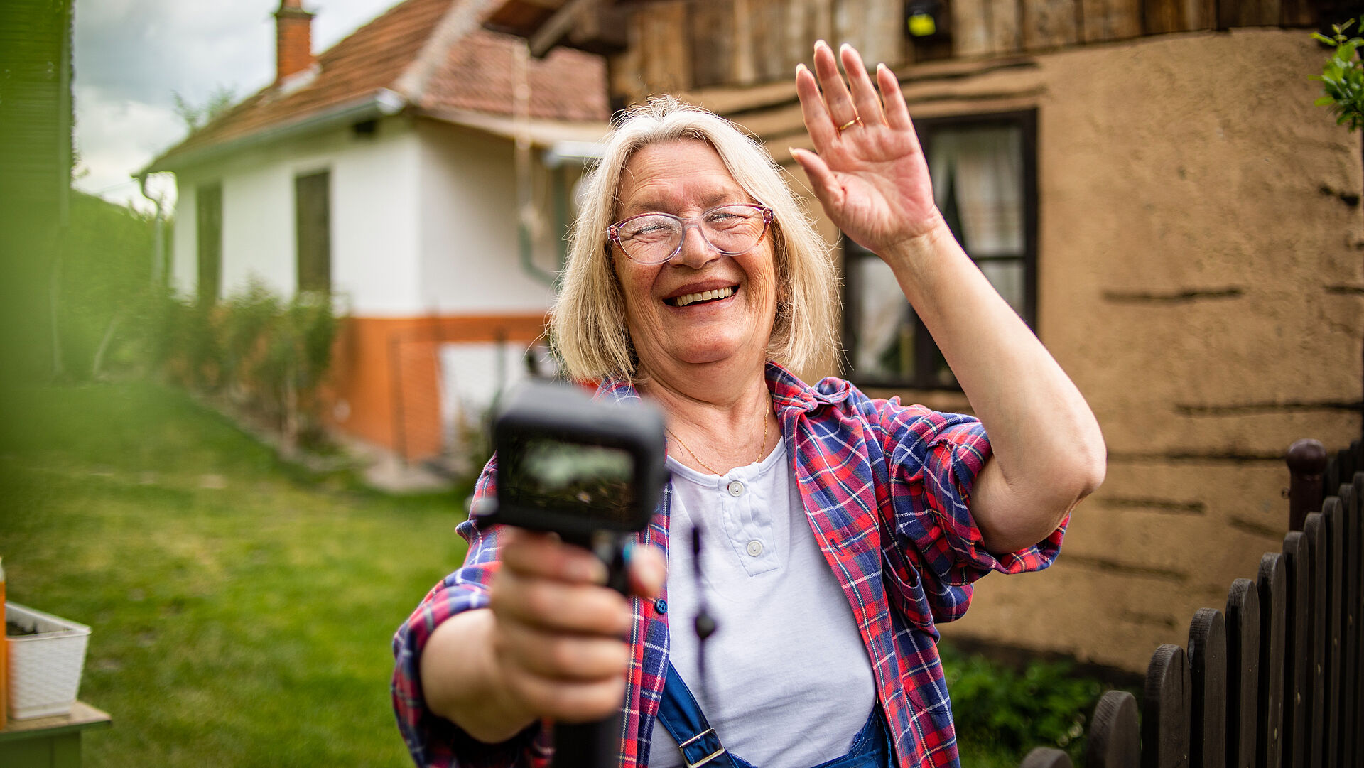 Eine Frau, die in ihrem Garten lachend in eine Kamera winkt.
