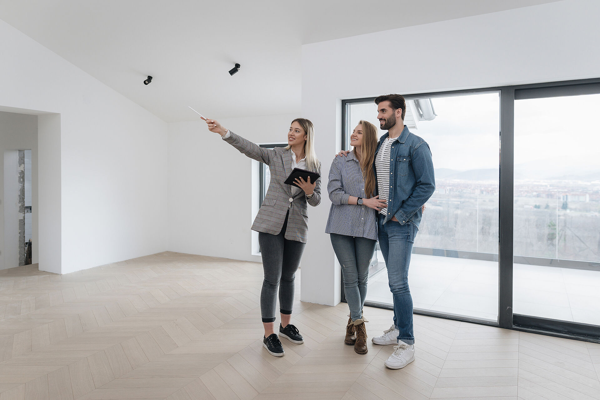 Eine Maklerin zeigt einem jungen Paar auf einem Tablet Daten zum Objekt. Alle drei stehen im Eingangsbereich einer leeren Wohnung.
