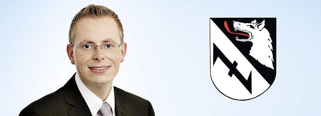 Herr Plapper vor blauem Hintergrund neben einem schwarz-weißen Wappen mit Wolf